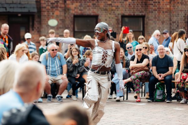 An image of a black male performer wearing a white top and trousers and a grey mask with a crowd watching behind him.