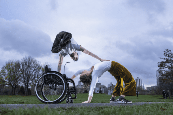 An image of a performer doing a handstand on a wheelchair with one hand holding the chair and the other leaning on a female performer who is doing a crab like pose. There is a cloudy sky in the background.