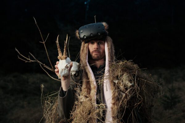 An image of a man dressed in nordic dress with a fluffy headdress holding an animal skull against a black background,