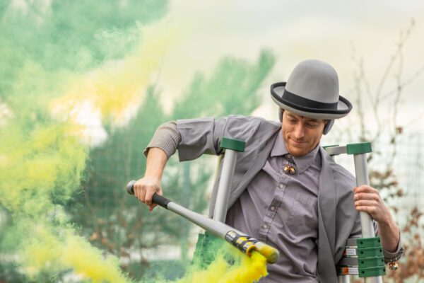 An image of a male performer wearing grey clothing and a top hat holding a flare that omits yellow smoke.