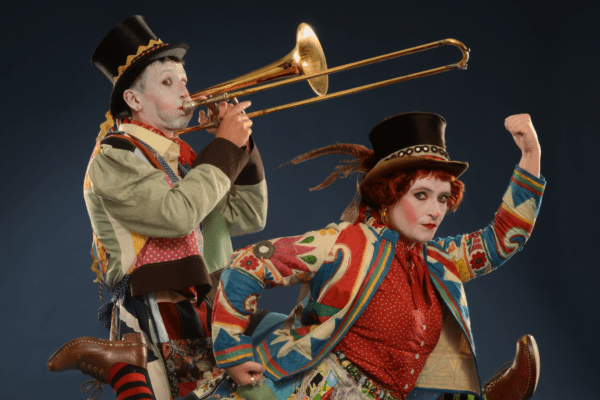 An image of two performers. The male performer is playing the trombone and the female is crouched down in front of him with her fist ib the iar. They are wearing colourful clothing and face paint against a black background.