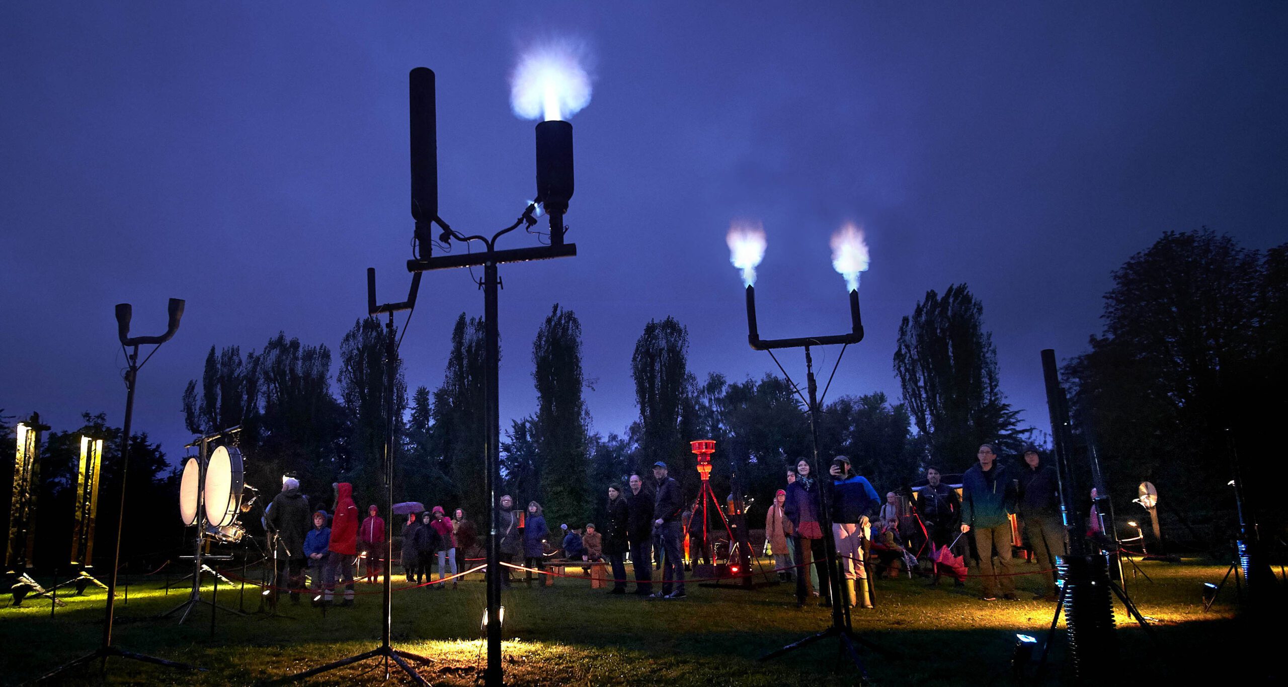 A night time image of a group of plinths fixed to the ground with prongs at the top omitting white light. There is a crowd and tall trees in the background.