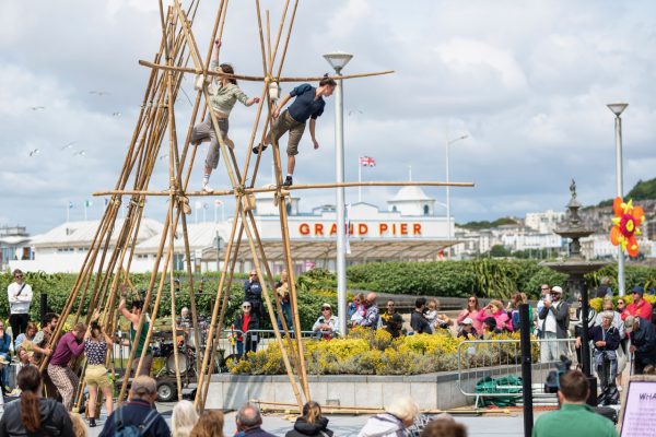 An image of a large bamboo structure with two performers weaving their way through it. There is a grand pier in the background and a crowd watching them below.