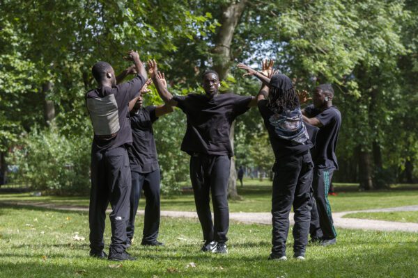An image of a group of performers dressed in black stood in a circle in a grassy area.