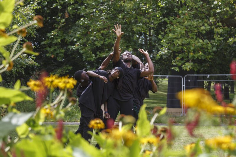 An image of a group of black male dancers intertwining and reaching upwards. They are all wearing black and there is greenery behind them.
