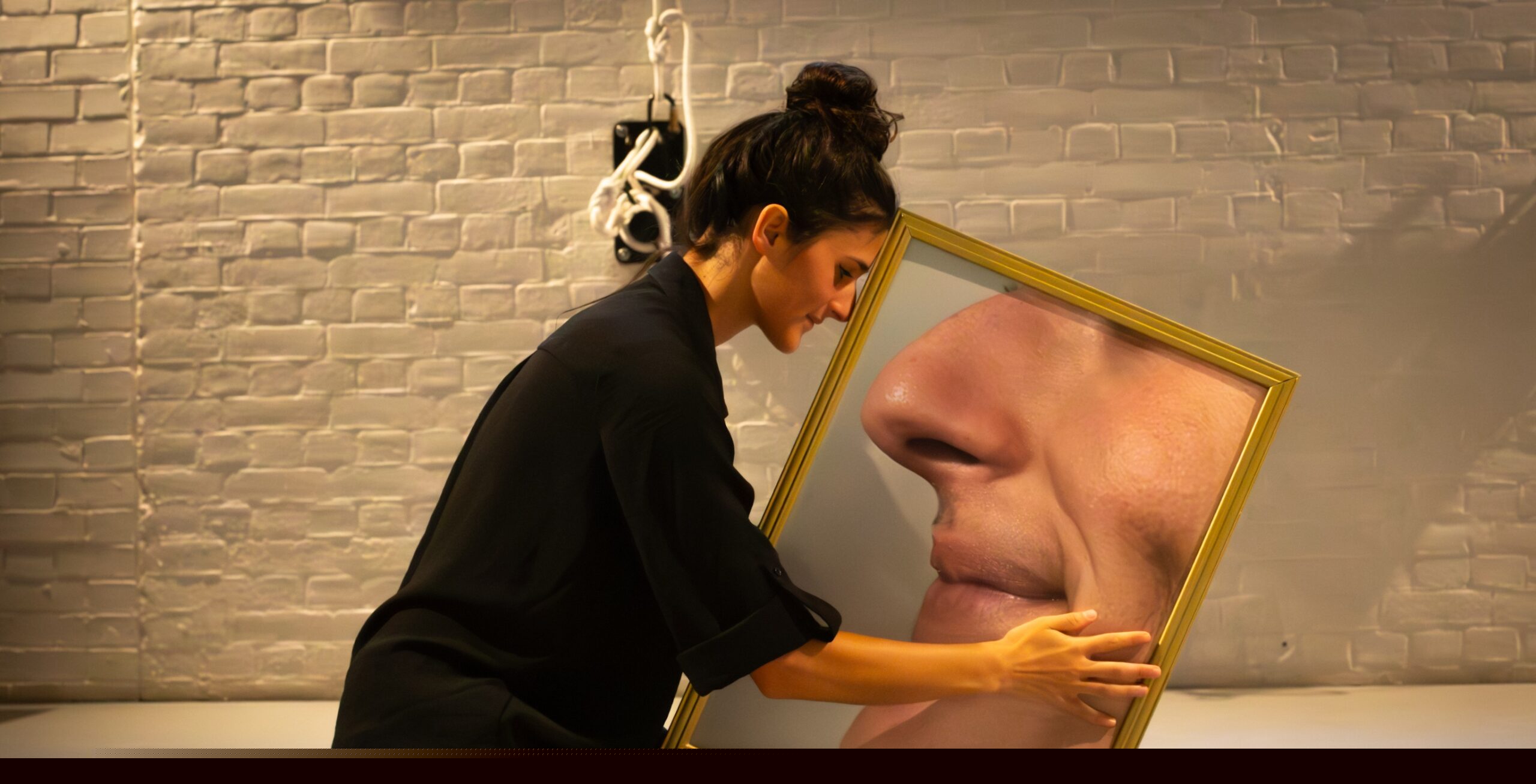 An image of a woman kneeling on the floor dressed in black. She is holding a painting of someone's face depicted sideways.
