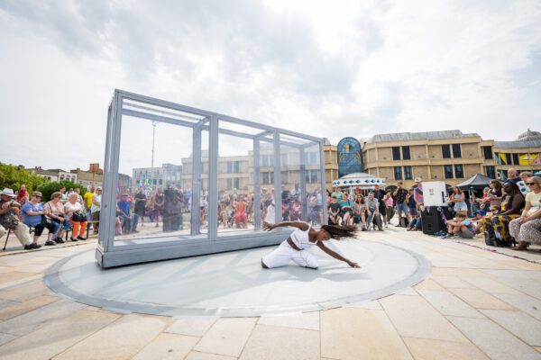 An image of a large transparent rectangular structure with a female dancer writhing on the floor beside it with a crowd in the background.