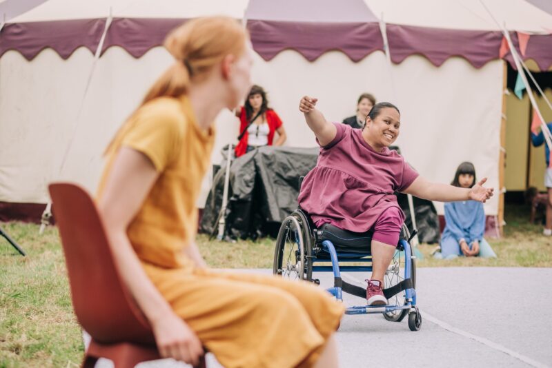 An image of a female performer who is a wheelchair user reaching out towards a woman sat on a chair, with a circus tent behind her.