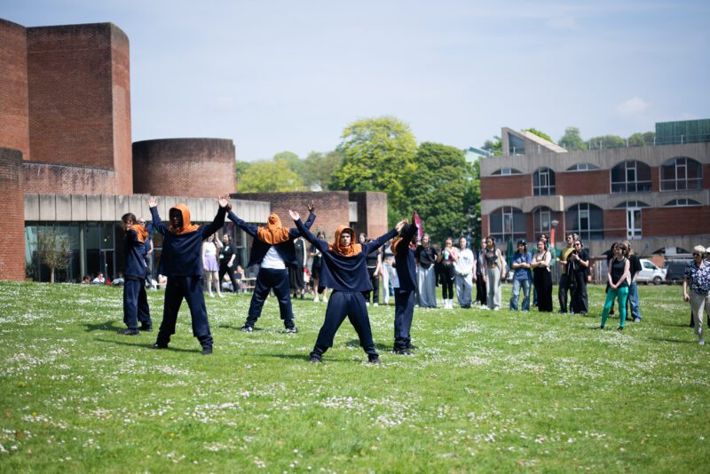 An image of a group of male dancers performing on a grassy surface. They are wearing dark blue and orange tracksuits and there is a small crowd behind them.