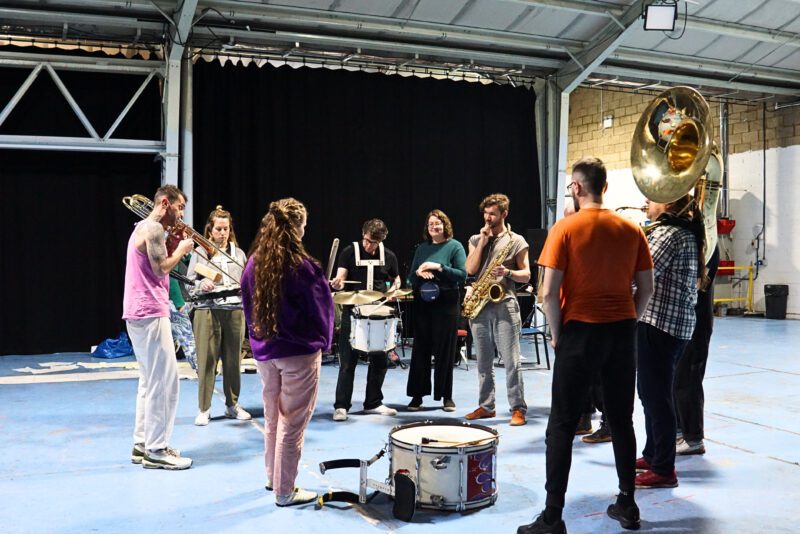 An image of a marching band stood in a circle in a rehearsal space playing various brass and percussion instruments