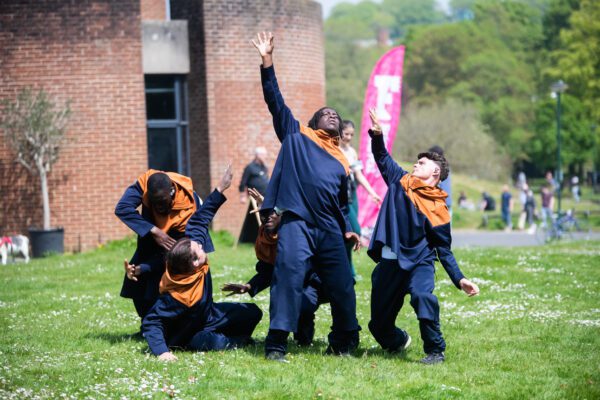 An image of a group of male dancers performing on a grassy surface. They are wearing dark blue and orange tracksuits.