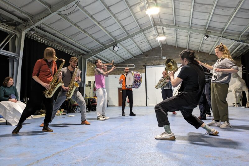 An image of a marching band stood in a circle in a rehearsal space playing various brass and percussion instruments