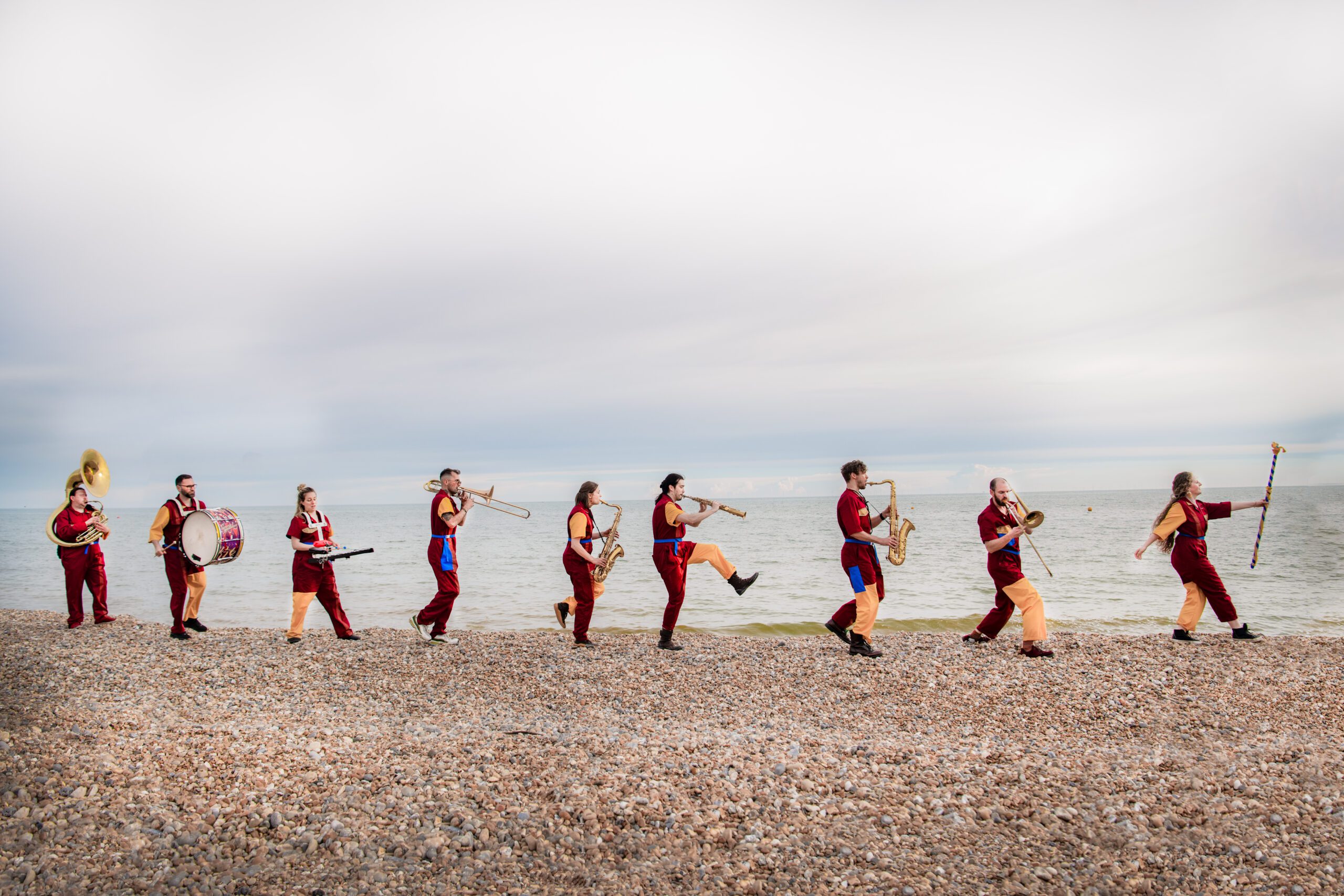 An image of a brass band walking along a beach playing various musical instruments. They are all wearing red and yellow clothing.