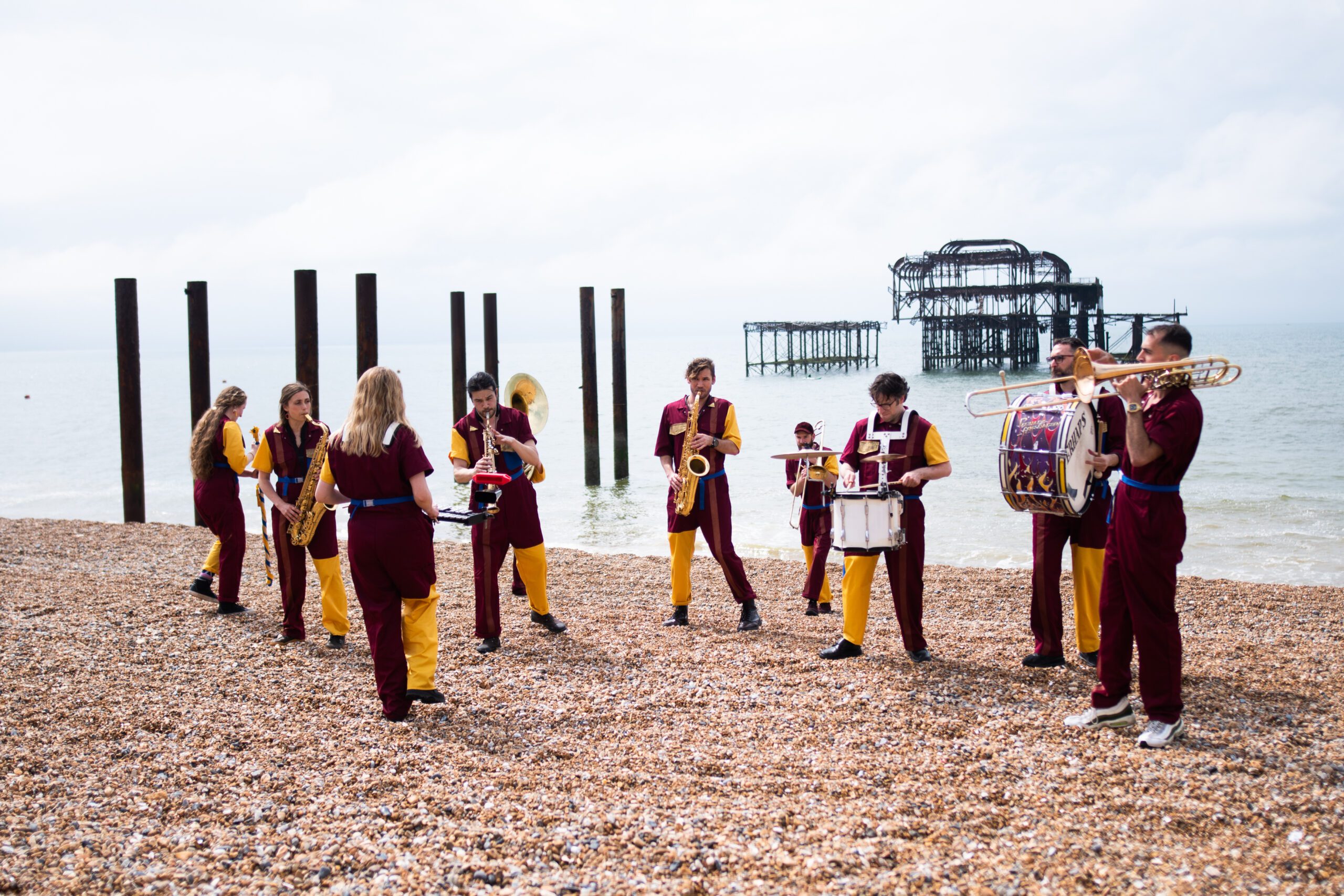 An image of a marching band stood on a beach with various brass and percussion instruments with a pier in the background.
