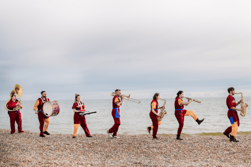 A brass band playing music while walking along a pebble beach against a cloudy sky scape.