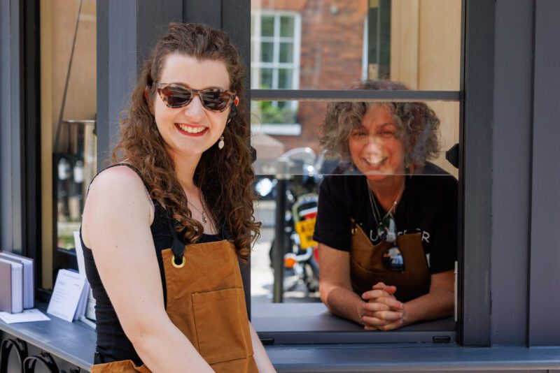 An image of a two women facing the camera and smiling. One is stood outside a kiosk wearing a brown apron and sunglasses. The other is stood inside with her elbows leaning on the counter and looking through the window.