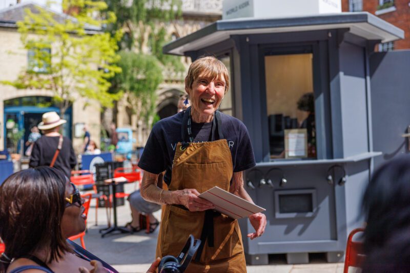An image of a person smiling at the camera wearing a brown apron with a dark t-shirt underneath, with a grey kiosk in the background.