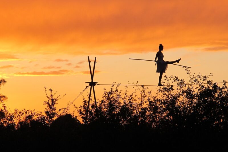 An image of a sunset orange sky with a silhouette of a tight rope walker in the foreground suspended above some bushes