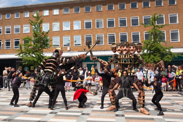 An image of a group of puppeteers and puppets performing a routine in a city square.