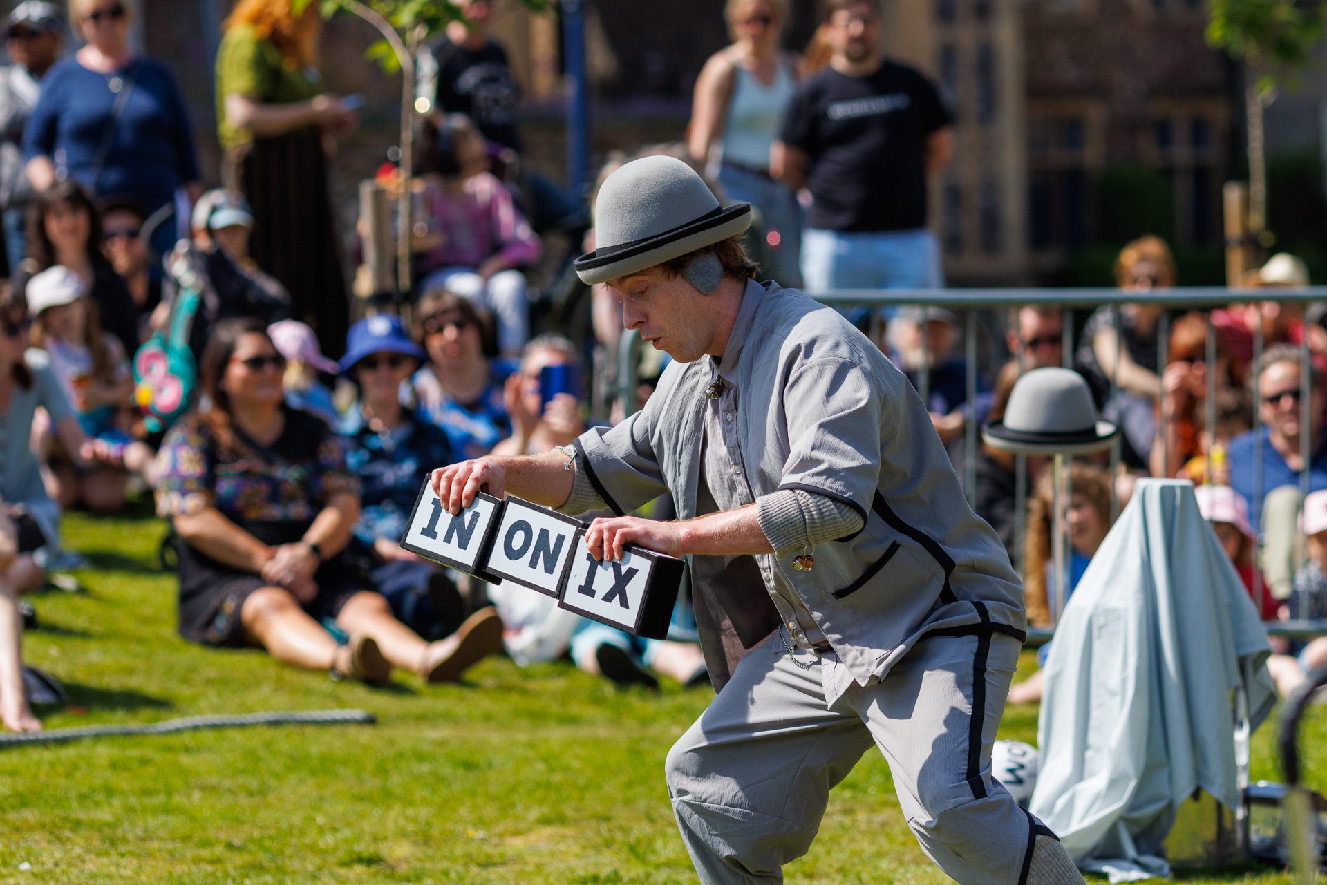 An image of a male performer wearing a grey hat, top and trousers. He is holding three rectangle shaped boxes with the letters IN ON IX written on them. There is a small crowd sat on the grass behind him.