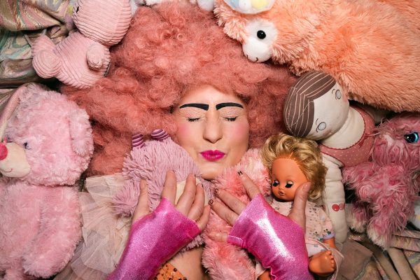 An image of a woman wearing a pink wig and gloves, surrounded by pink toys and teddy bears.