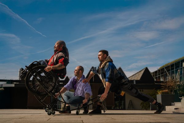 An image of three male dancers against a blue sky backdrop. The principal dancer is a wheelchair user and to the left of the image.
