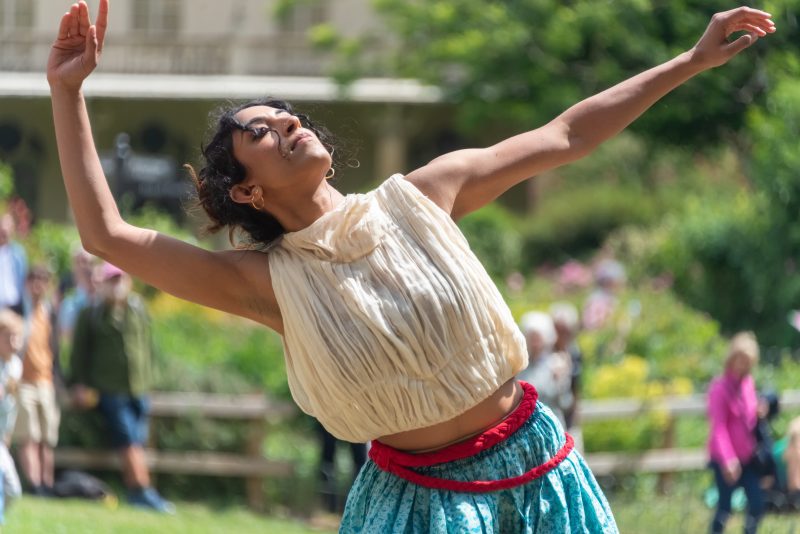 An image of a female dancer wearing a cream sleeveless top and green skirt.