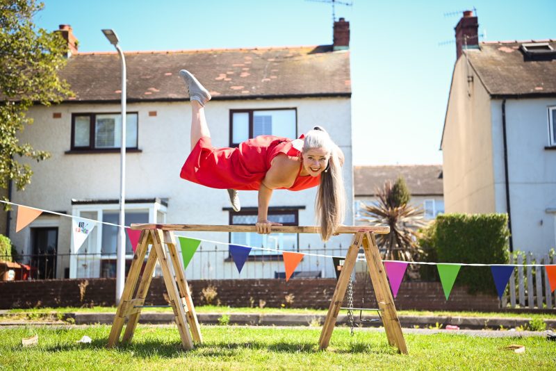 An image of a performer suspended between two wooden plinths on a grassy area with a semi detached house in the background. She is wearing a red playsuit.