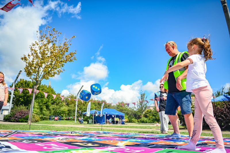 An image of a festival volunteer and a child walking across a colourful floor in a park setting, with blue sky and trees in the background.