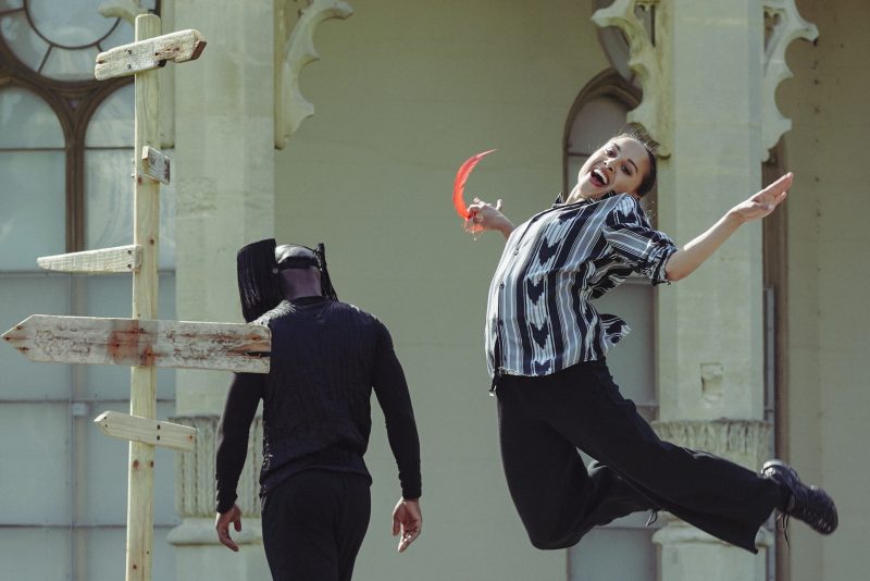 An image of two performance artists doing street art with a cream coloured building in the background. One female artist is jumping in the air and the other is wearing all black and walking away from her.