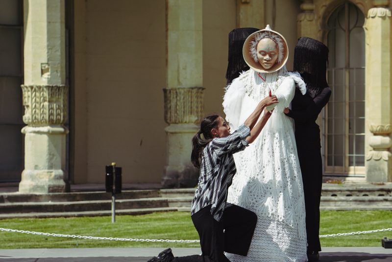 Two people assisting with a masked figure wearing a long white dress, with a historic building in the background.