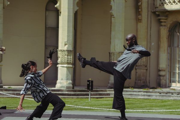 An image of two people performing street art. One person is kicking towards the other person, and the other person is holding their hand up and bending backwards.