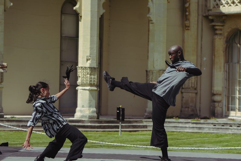 An image of two people performing street art. One person is kicking towards the other person, and the other person is holding their hand up and bending backwards.