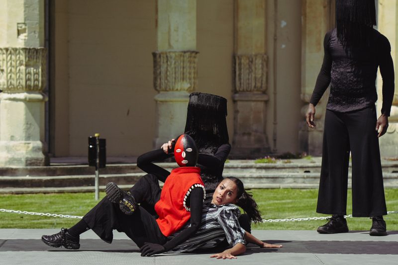 An image of am outdoor theatre performance with three people wearing black and red. Two people are lying on the floor with one wearing a mask and the other looking at the camera. There is a hooded figure stood up watching over them.
