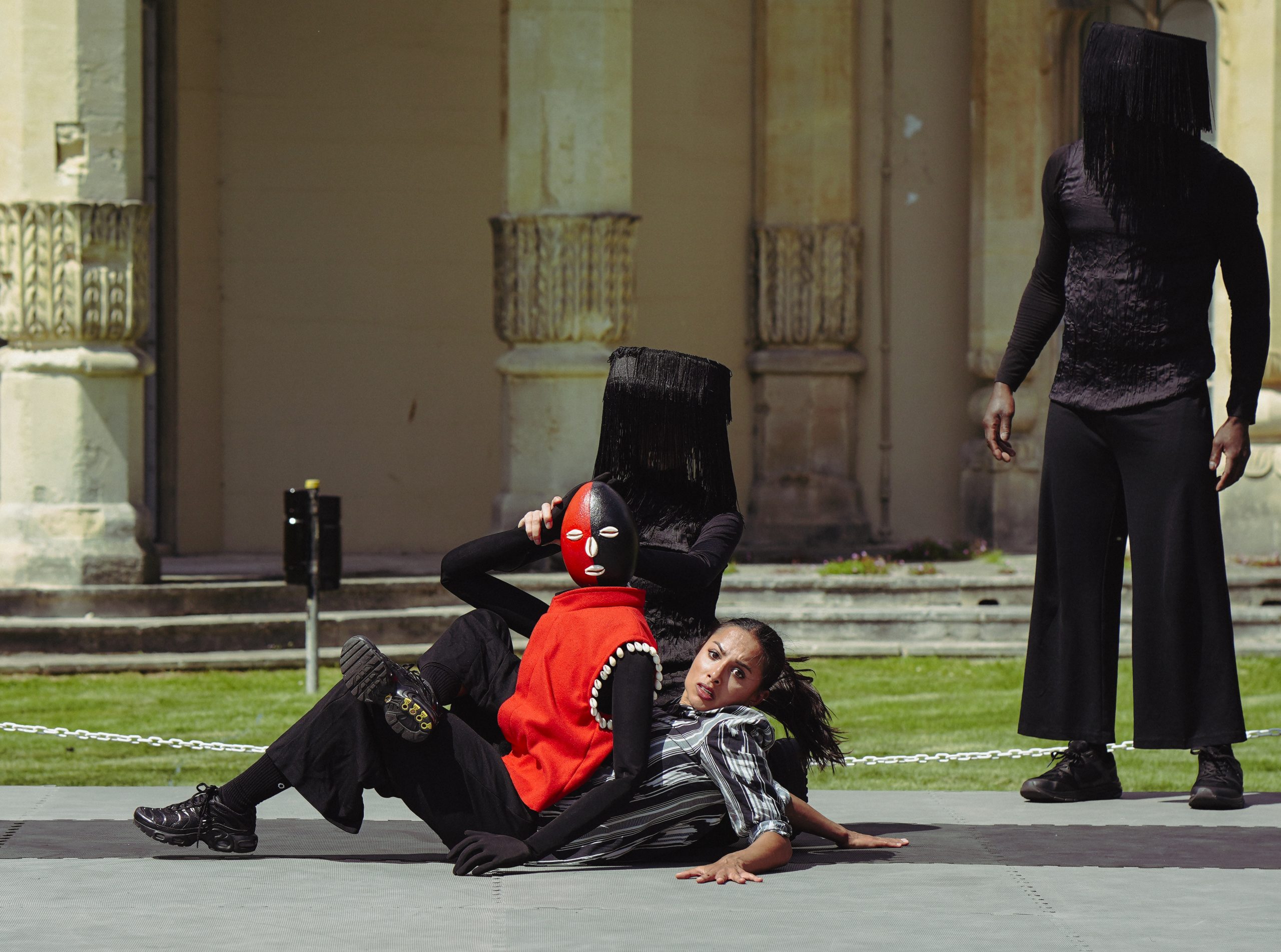 An image of am outdoor theatre performance with three people wearing black and red. Two people are lying on the floor with one wearing a mask and the other looking at the camera. There is a hooded figure stood up watching over them.