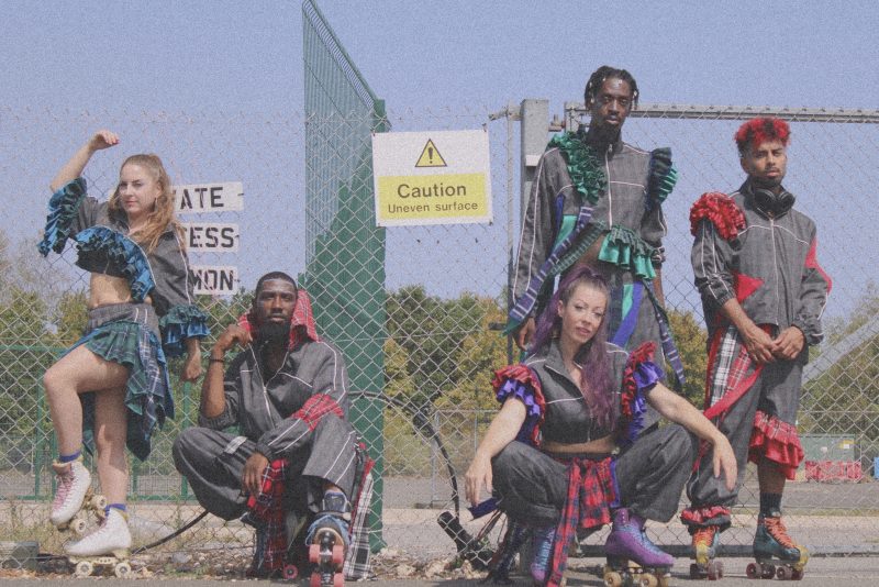 An image of 5 dance artists wearing tartan and grey outfits and roller skates stood outside a fence with a sign saying 'caution uneven surface'.