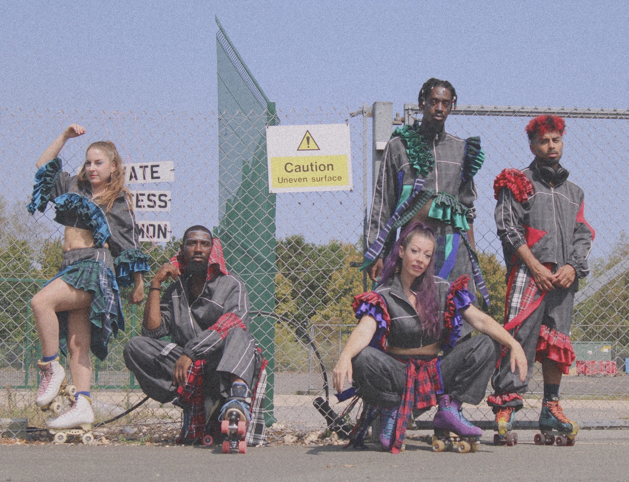 An image of 5 dance artists wearing tartan and grey outfits and roller skates stood outside a fence with a sign saying 'caution uneven surface'.