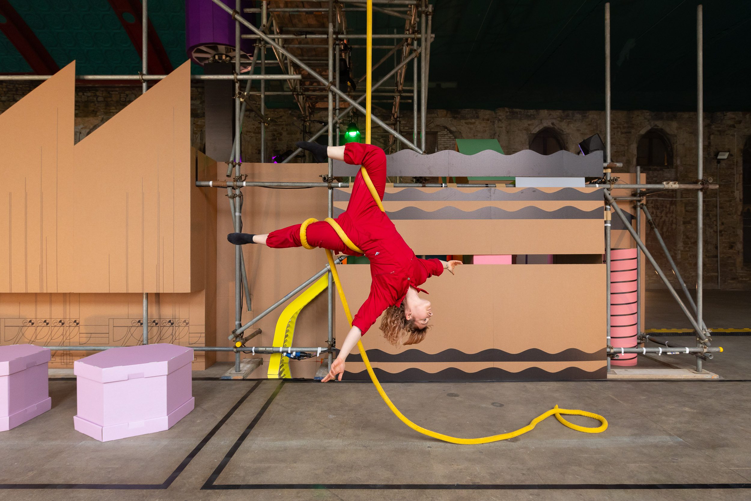 A circus performer hanging upside down from a yellow rope wearing a red boiler suit, with a sparse background.