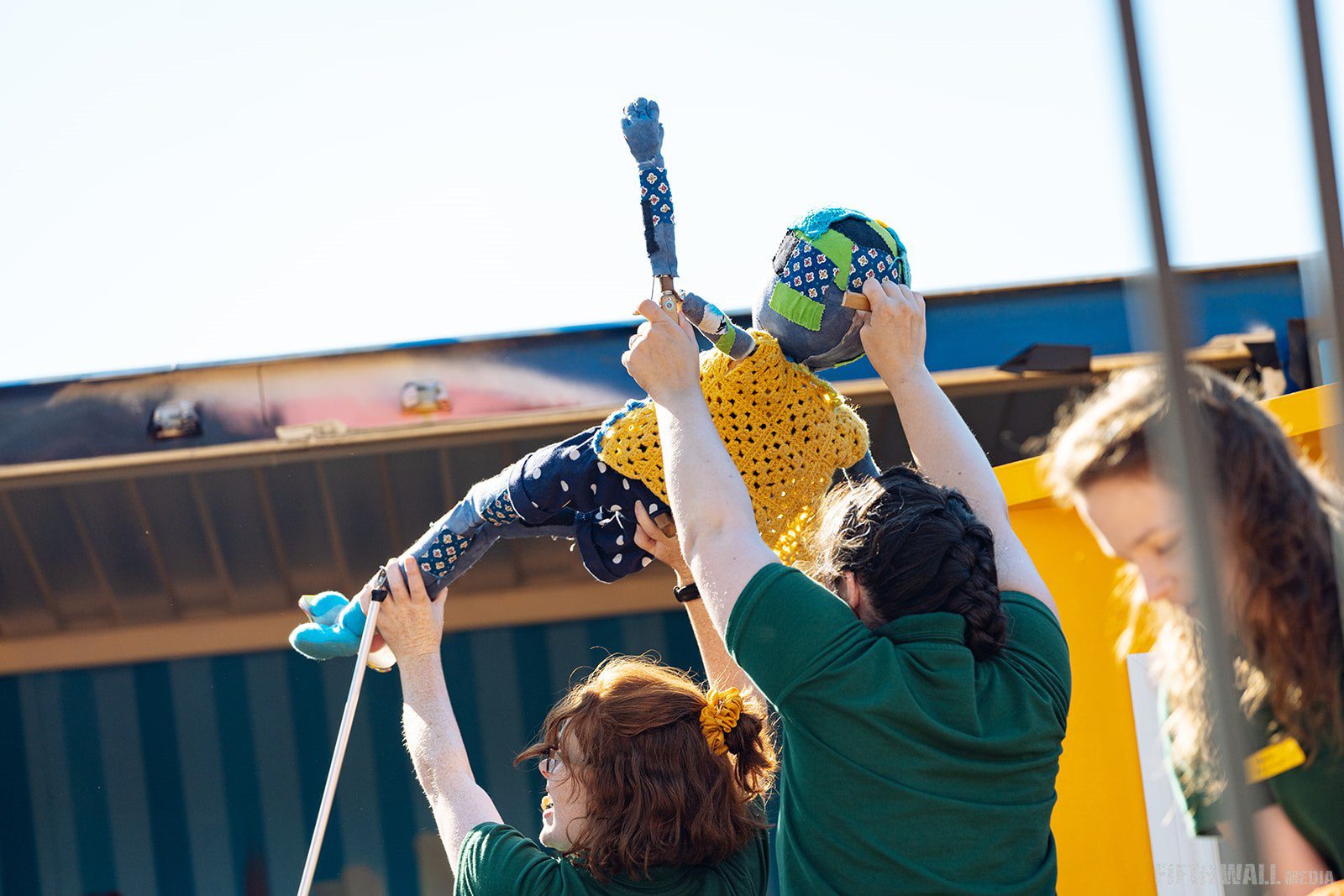 Two people lifting up a child puppet who is wearing yellow.