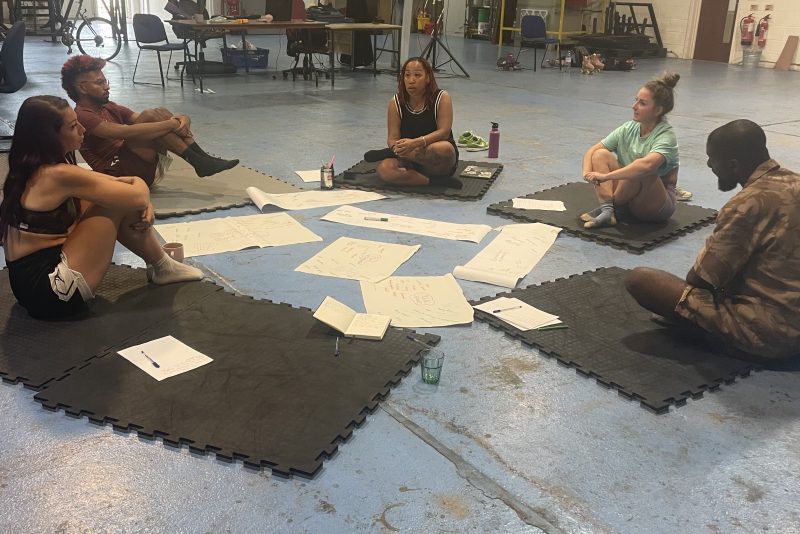An image of 5 people, 3 female and 2 male, sat in a circle on black mats on the floor in a dance studio, surrounded by white papers