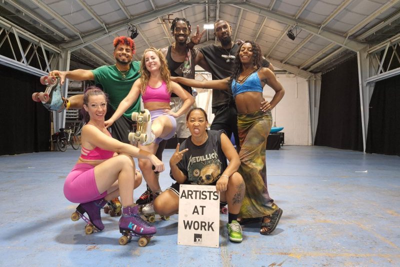 An image of a group of roller skate dancers posing for the camera in a large empty warehouse. There are 4 females and 3 males, all wearing casual and sports clothing. A dancer at the front is crouched down holding a sign saying 'artists at work'.