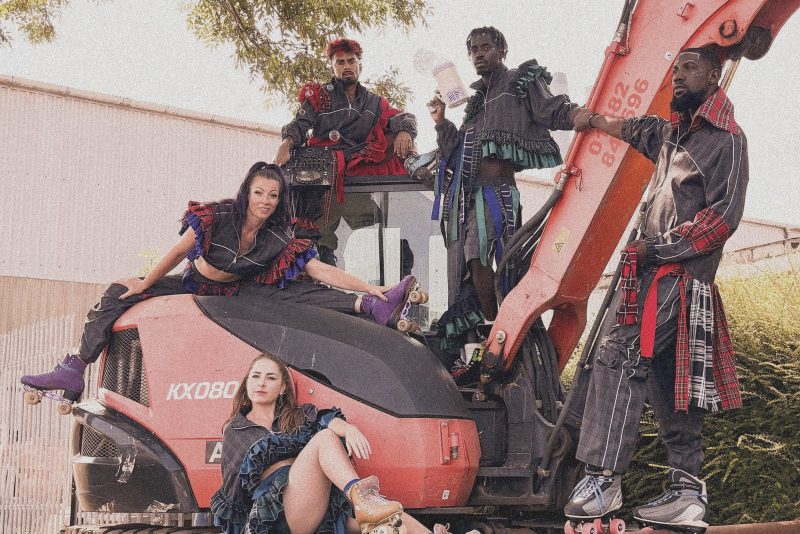 An image of 5 skate dancers, 4 male and 2 female, sat and stood around a large red digger. Without Walls is a network of 35 festivals who commission and co-create outdoor art for everyone across the UK.