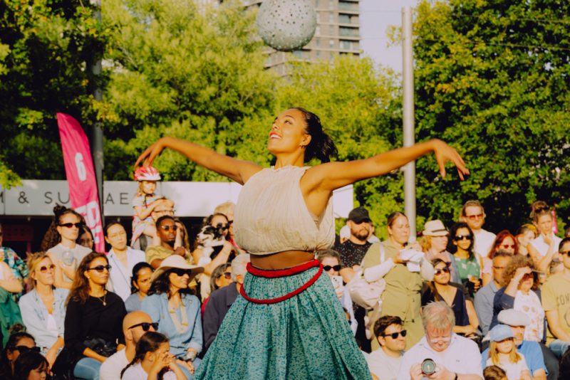 An image of a female dancer with her arms outstretched wearing traditional Indian dress with a crowd behind her.