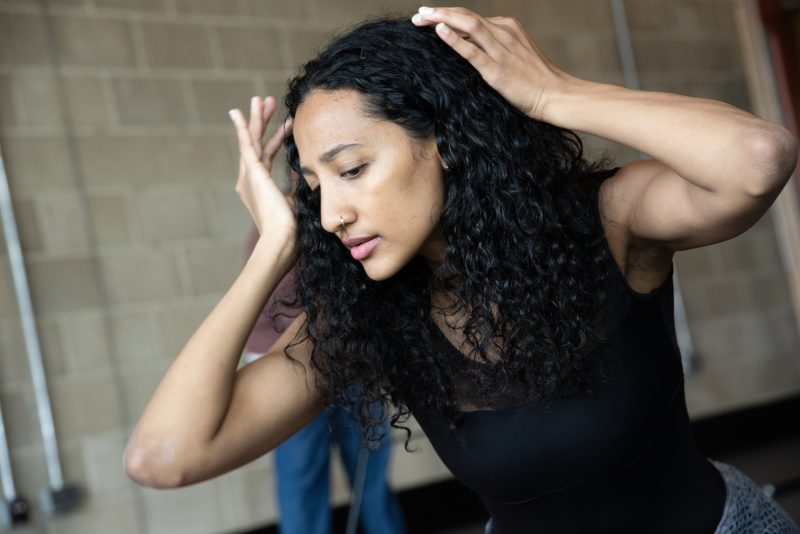 An image of a female dancer wearing a dark blue vest and long dark curly hair, with her arms lifted and her hands on her head.