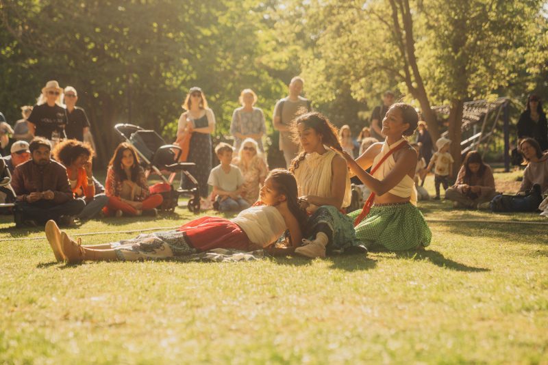 An image of three female dancers sat and lying on grass with a sunlit backdrop and captivated audience.