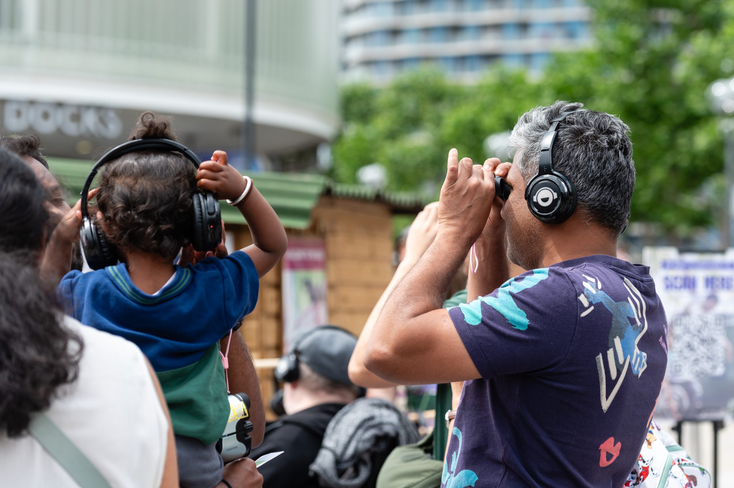 An image of a man and a young child wearing headphones. The man is looking through binoculars and wearing a grey T-shirt.
