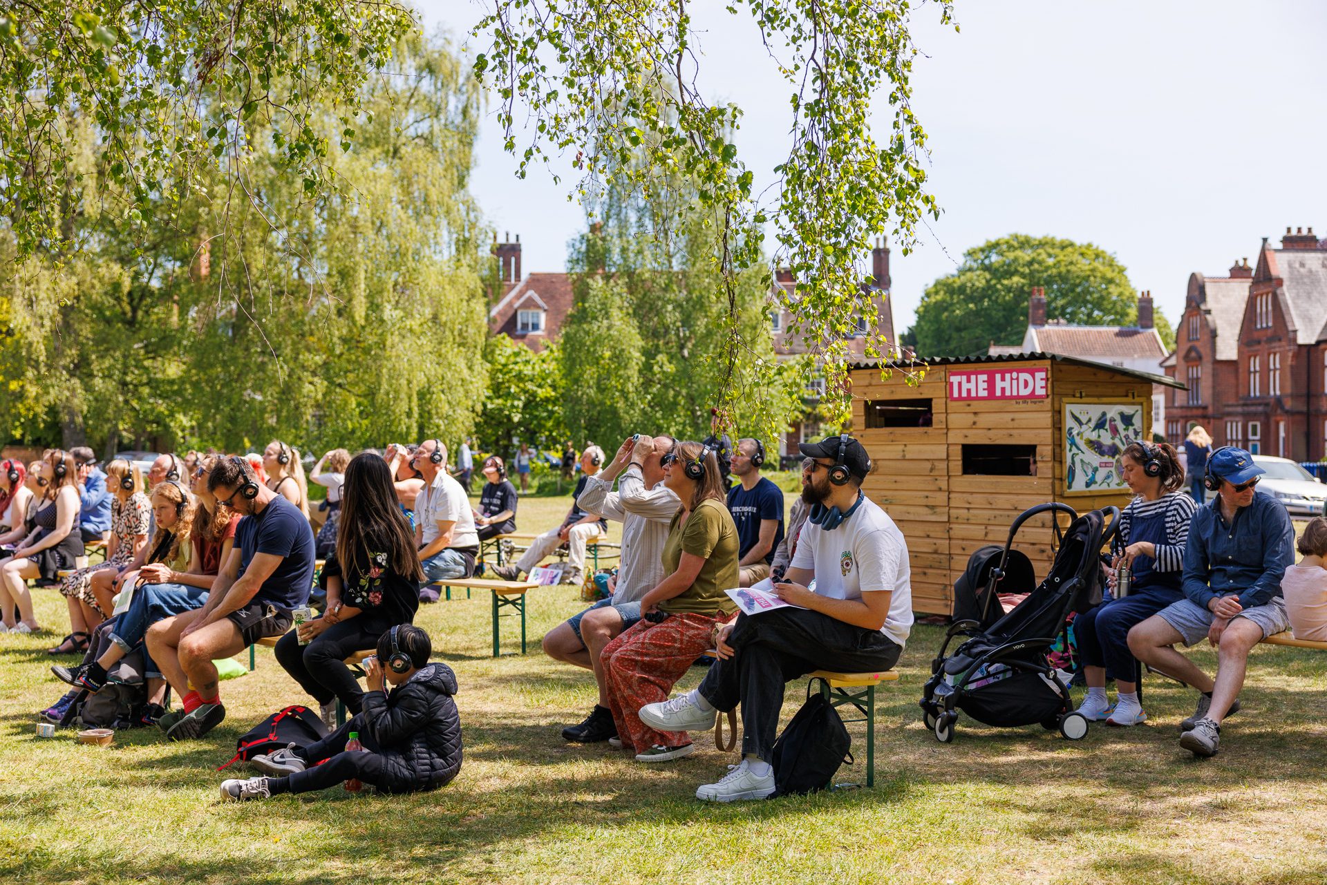 An image of a group of people sat on chairs looking towards the left wearing headphones. Some people are looking through headphones. There is a shed behind them with 'The Hide' inscribed on the top left.