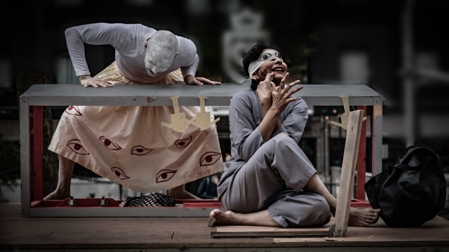 An image of two dancers, with one leaning over a long grey table. The dancer in the foreground is wearing a blind fold.