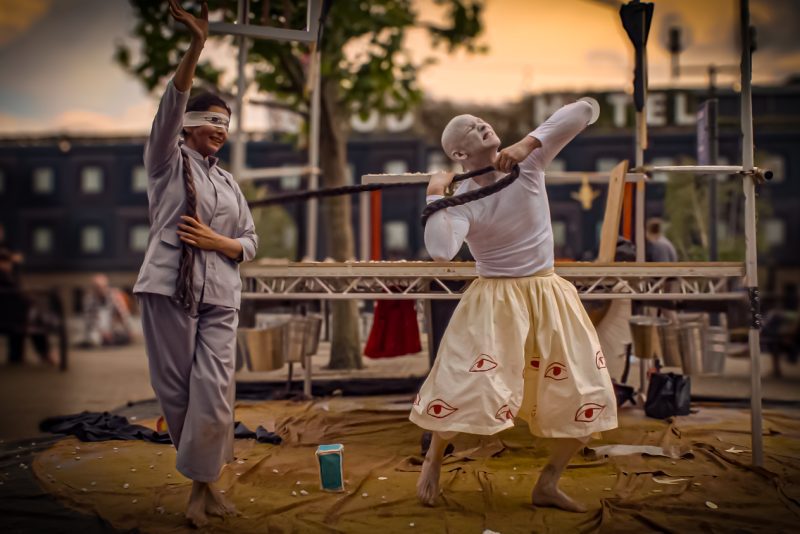 An image of 2 dancers, with one pulling a rope from behind the other. They are wearing grey and cream clothing, with a rigged platform in the background with silver hanging buckets.