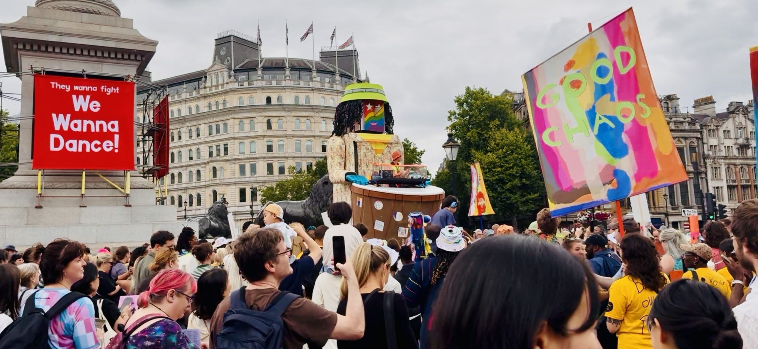 A street crowd gathering around a large puppet. There is a rad square banner tied to a monument in the background with the caption 'We Wanna Dance!'.